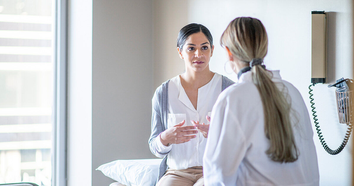 A patient sits on an examination table while speaking with a healthcare professional in a clinical setting. The room is well-lit with natural light coming from a window. Medical equipment is visible on the wall, and both individuals appear engaged in conversation. The scene conveys a calm and professional atmosphere.
