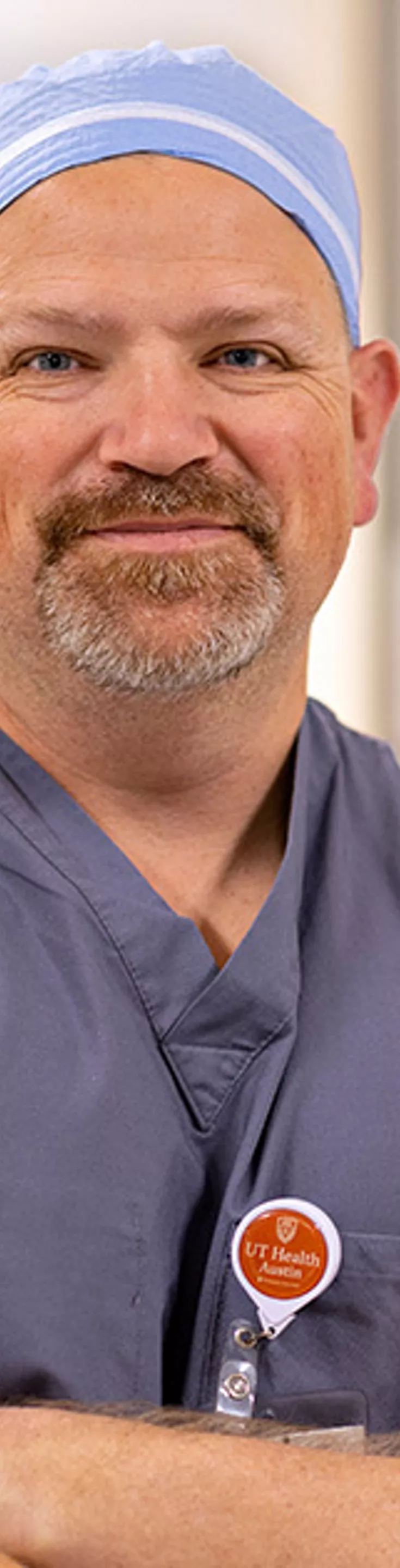 A person wearing blue medical scrubs and a cap stands with arms crossed in a brightly lit hospital hallway. The individual has a visible ID badge with orange and white elements. The background features soft, neutral tones and blurred doors, suggesting a clinical environment. The overall mood is professional and calm.
