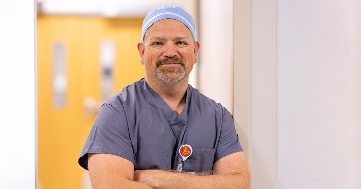A person wearing blue medical scrubs and a cap stands with arms crossed in a brightly lit hospital hallway. The individual has a visible ID badge with orange and white elements. The background features soft, neutral tones and blurred doors, suggesting a clinical environment. The overall mood is professional and calm.