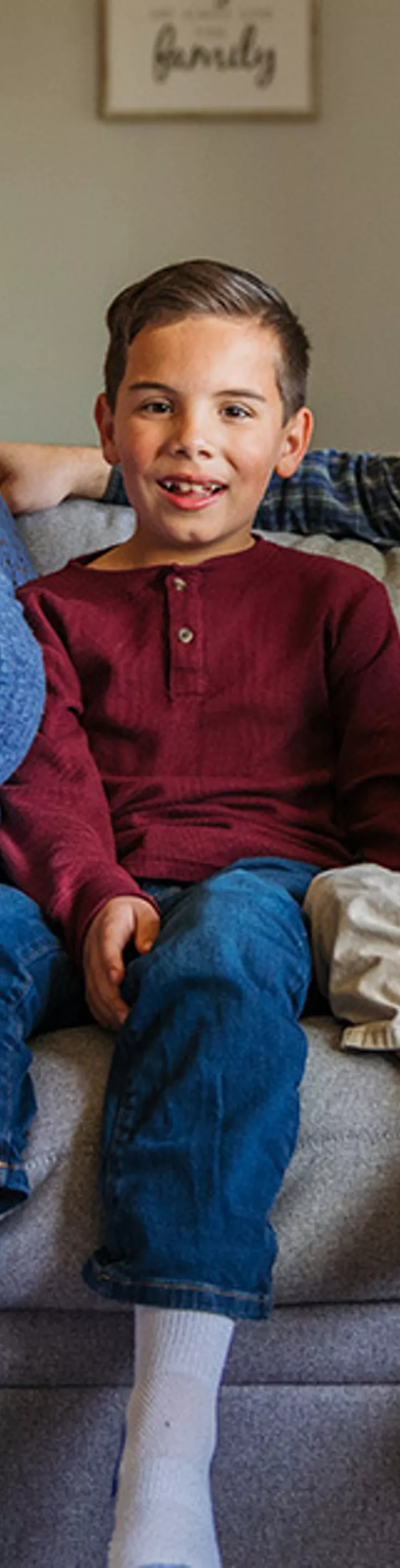 A family of four, including two adults and two children, sits closely together on a light gray sofa in a modern living room. The adults are dressed in casual attire, while the children wear colorful tops and socks. The setting is warm and inviting, with neutral tones and soft natural light. A wall sign reading 'family' is visible in the background, enhancing the cozy atmosphere.