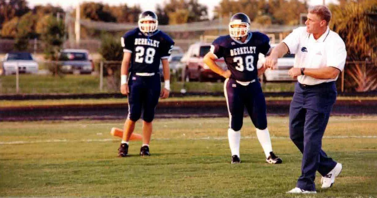 Two football players in navy uniforms with visible numbers '28' and '38' stand on a grassy field, accompanied by a coach giving instructions. The players' jerseys display the word 'BERKELEY' in large letters. The background includes parked cars and trees, suggesting an outdoor sports facility. The scene captures a moment of teamwork and preparation.