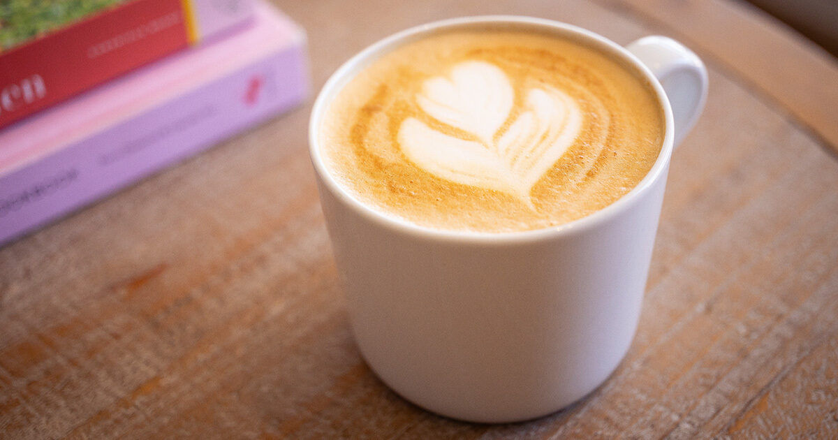 A white ceramic mug filled with a latte featuring heart-shaped foam art sits on a rustic wooden table. In the background, two pastel-colored books are partially visible, adding a cozy and relaxed atmosphere. The image highlights the creamy texture and artistic presentation of the coffee. No visible numeric values or identifiable brands are present.