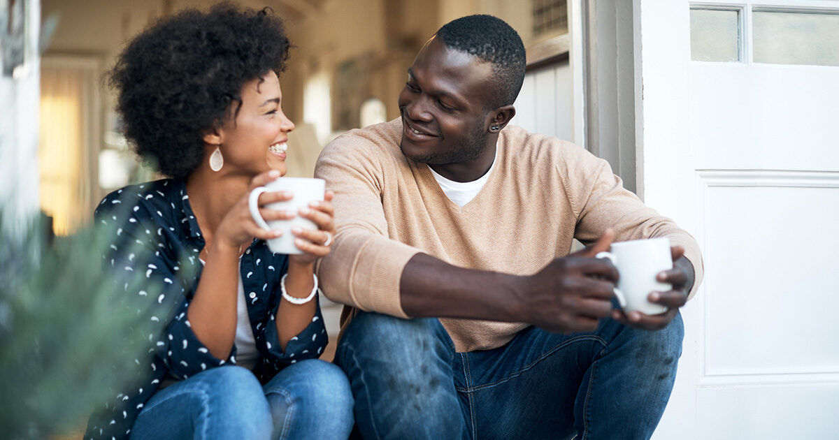 Two adults sit closely on outdoor steps, each holding a white mug. The setting appears relaxed and homey, with natural daylight and casual attire. The scene conveys a warm, intimate moment, with soft focus on the background and neutral color tones.