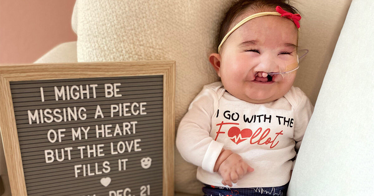 An infant sits comfortably on a soft chair next to a letter board displaying a sentimental message. The child wears a white shirt with red and black text, featuring a heart graphic. The setting is warm and inviting, with neutral tones and plush textures. The sign includes the phrase 'I MIGHT BE MISSING A PIECE OF MY HEART BUT THE LOVE FILLS IT' and 'DEC. 21' as visible numeric text.