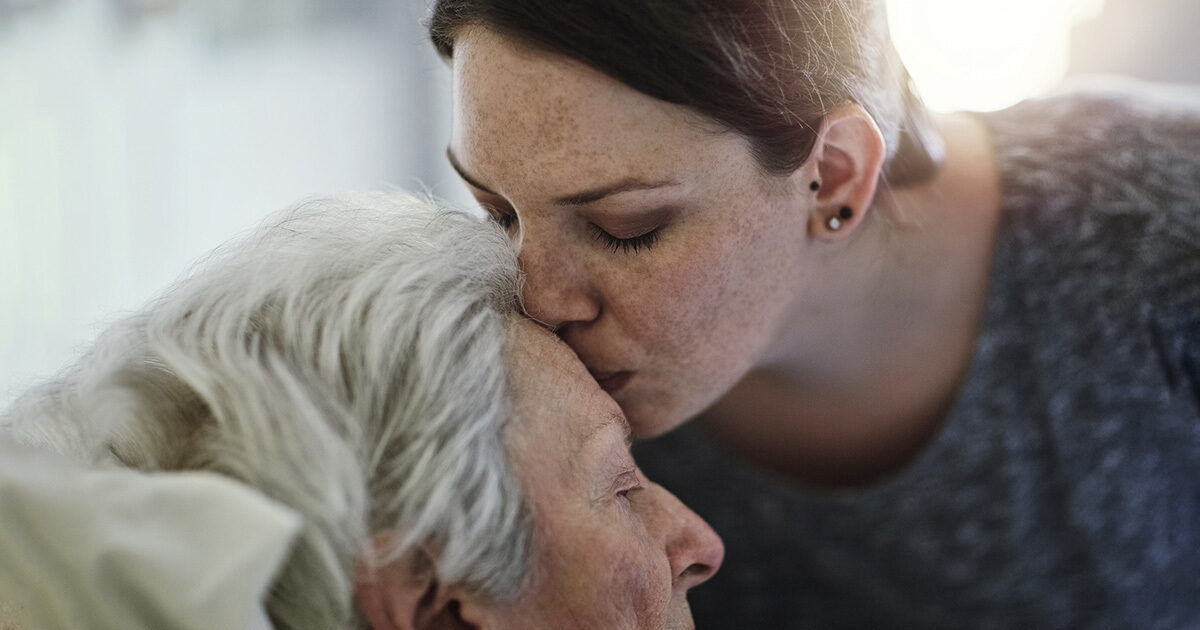 A young person with short hair and an earring leans in close to an elderly individual with white hair. The setting appears to be indoors, with soft natural light filtering in from the background. The mood is gentle and caring, suggesting a moment of support or empathy. No visible text or numbers are present in the image.