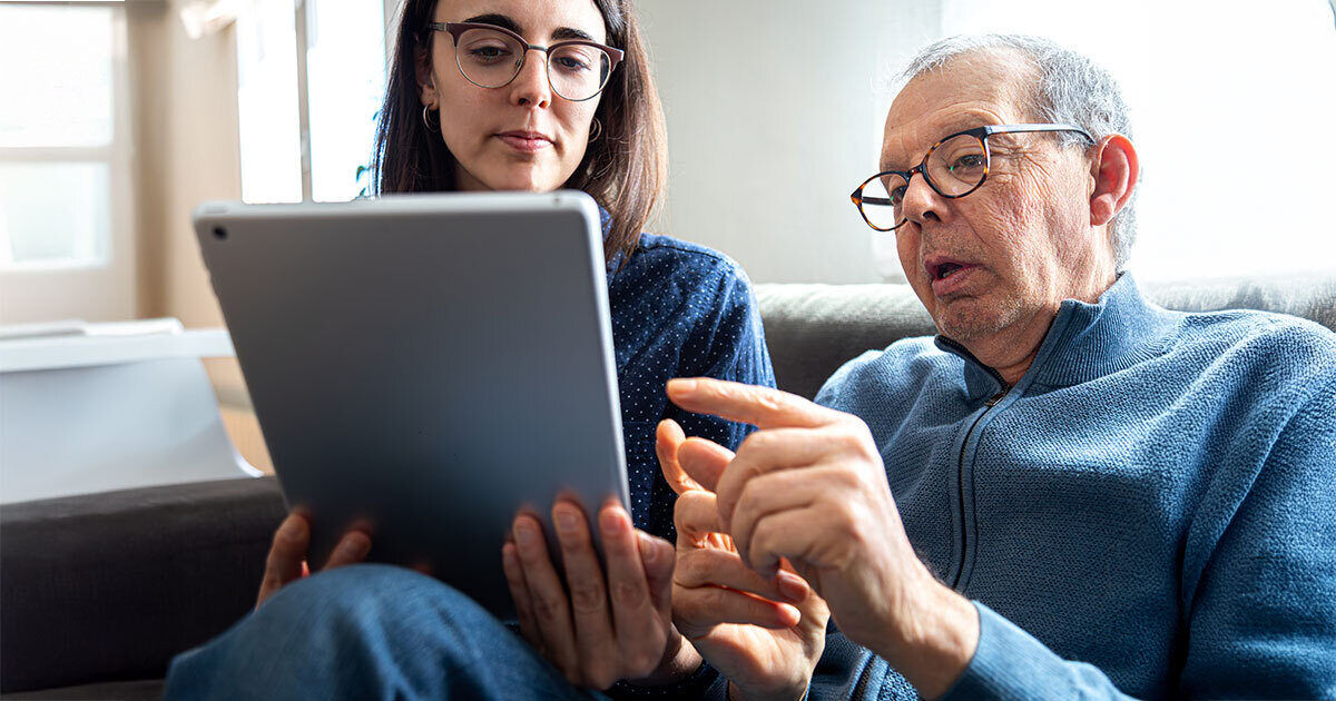 An older man and a woman are seated together on a couch, engaging with a large tablet device. The setting appears to be a cozy living room with soft natural lighting. The man is pointing at the tablet screen while the woman holds it, suggesting a collaborative or instructional activity. The overall mood is relaxed and focused, with a neutral color palette.