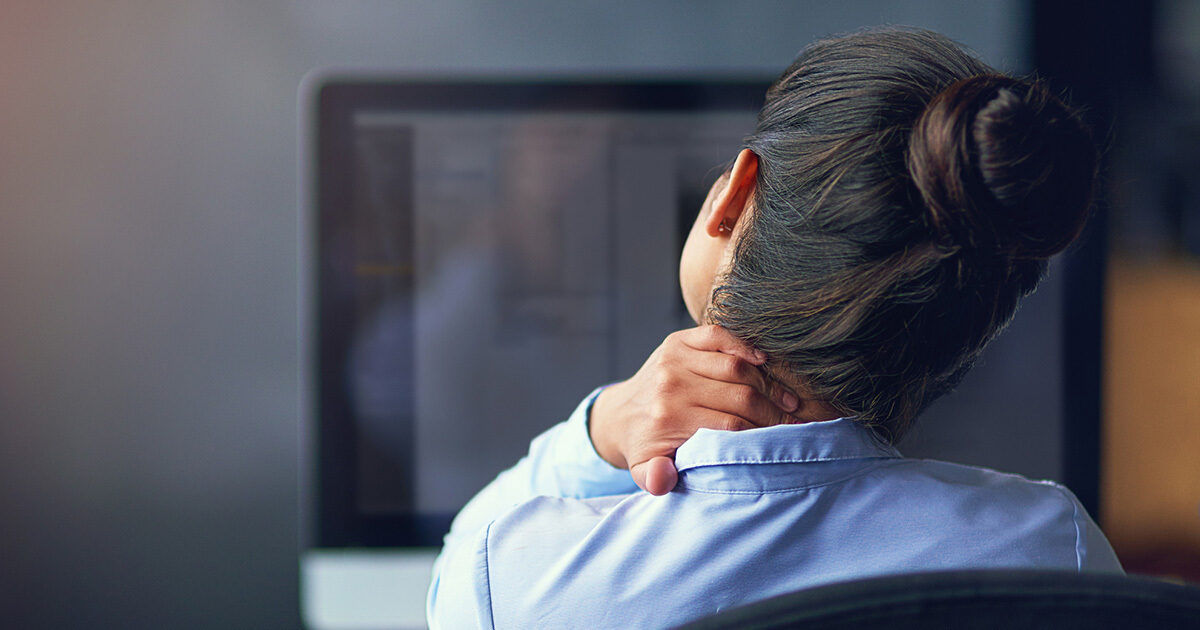 A woman sits at a desk in front of a computer monitor, holding her neck as if experiencing discomfort or pain. The setting appears to be an office environment with a blurred background, suggesting a workday scene. She wears a light blue shirt and her hair is tied in a neat bun. The image conveys a mood of fatigue or strain, commonly associated with prolonged desk work.