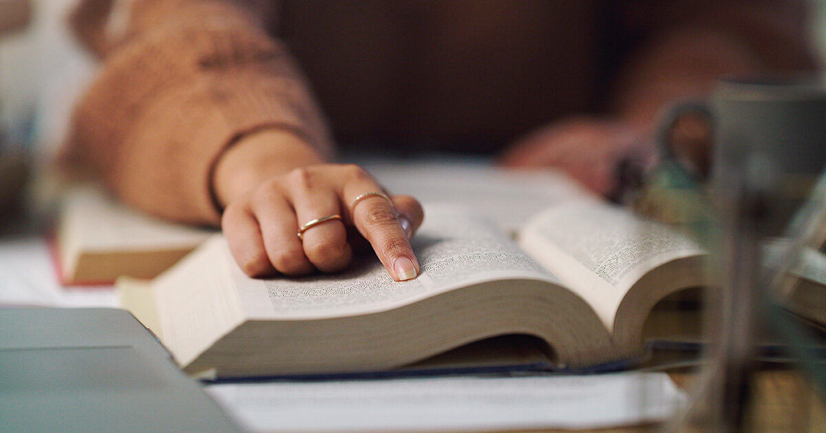 A close-up view shows a personâ€™s hand pointing at text in an open book, suggesting focused reading or studying. The individual appears to be wearing a brown sweater and a simple ring. The setting includes a blurred background with papers and a mug, indicating a study or work environment. The overall mood is calm and studious, with warm, neutral tones.