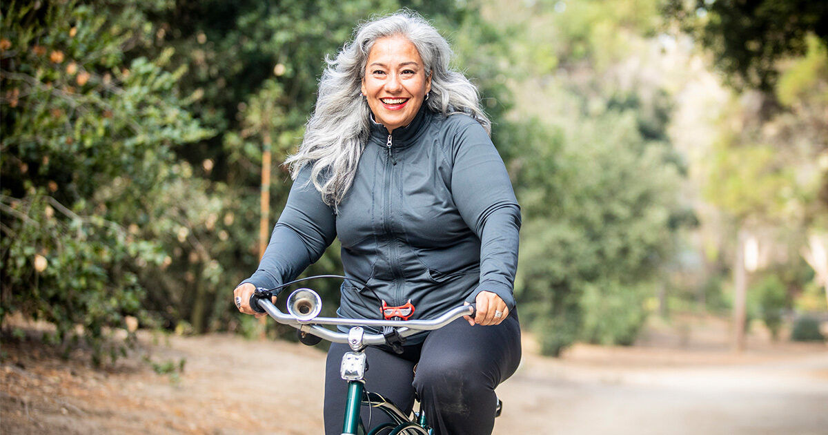 An older woman with long gray hair rides a bicycle along a dirt path in a wooded park. She is dressed in a dark athletic jacket and pants, enjoying a leisurely ride surrounded by greenery. The setting is natural and peaceful, with soft daylight filtering through the trees. The overall mood is relaxed and active, emphasizing outdoor recreation.