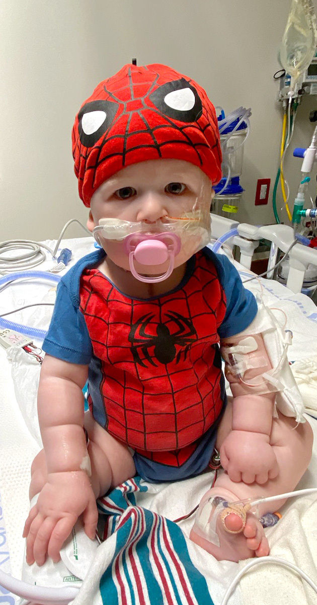 A young child is dressed in a red and blue superhero-themed outfit with a matching hat, sitting upright on a hospital bed. Medical equipment and IV lines are visible, indicating a healthcare setting. The child is surrounded by clinical devices and a striped hospital blanket. The environment is brightly lit, with a focus on the child's costume and medical care context.