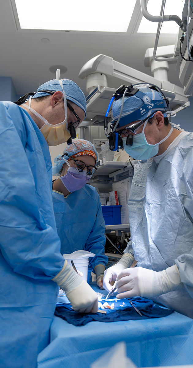 A team of surgeons is actively engaged in a surgical procedure in a well-equipped operating room. The medical professionals are wearing blue surgical gowns, gloves, and caps, with one using a headlamp for enhanced visibility. The environment is sterile and clinical, with advanced medical equipment visible in the background. The overall mood is focused and professional, highlighting teamwork and precision.