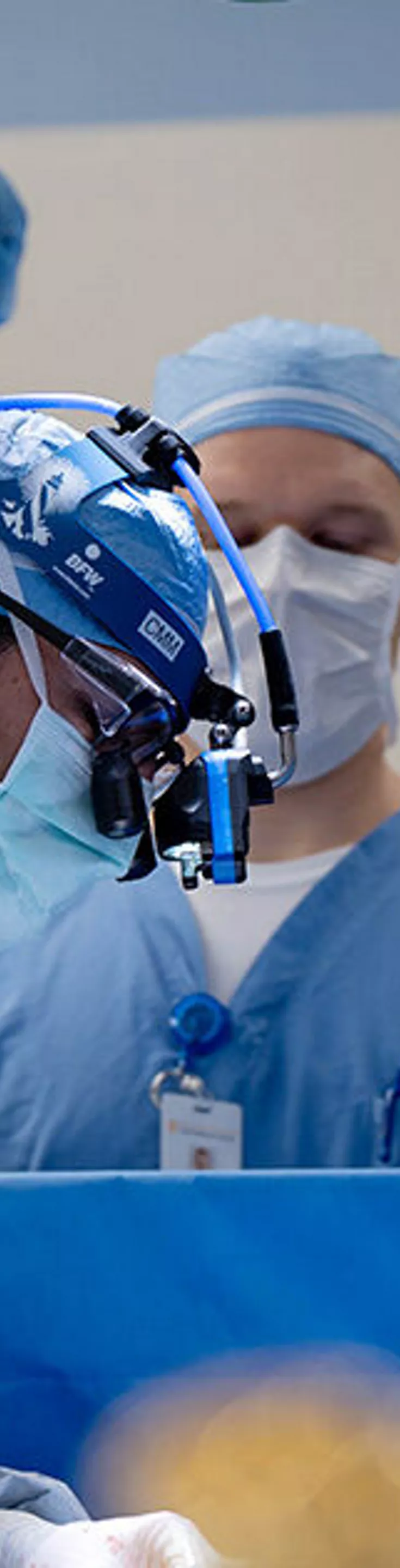 A group of healthcare professionals is gathered around an operating table, actively engaged in a surgical procedure. The setting is a modern hospital operating room, with all team members wearing blue surgical gowns, masks, and caps. Medical equipment and monitors are visible in the background, emphasizing a sterile and clinical environment. The image conveys focus, teamwork, and a professional medical atmosphere.