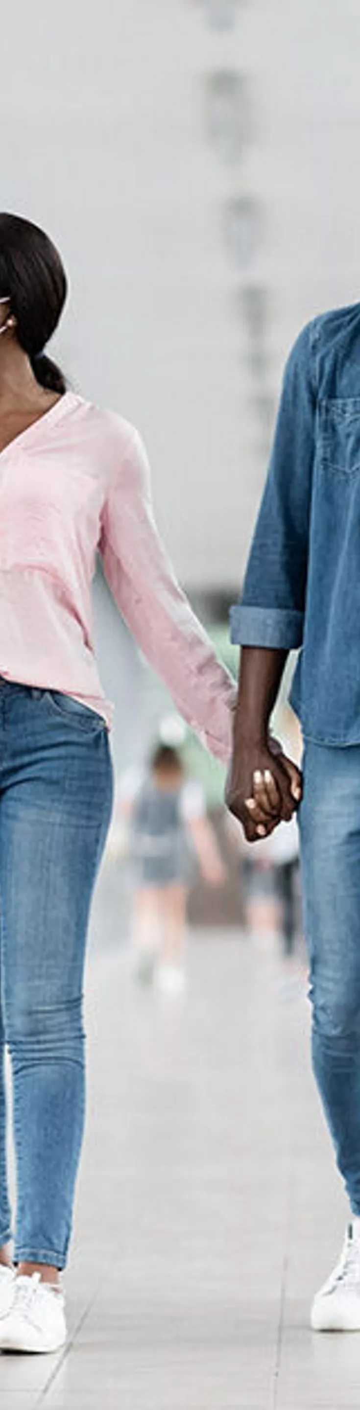 A young couple is seen walking side by side through a modern airport terminal, each pulling a rolling suitcase. Both individuals are casually dressed and wearing face masks, suggesting a travel scenario during a health-conscious period. The setting features bright natural light, glass walls, and a spacious corridor, emphasizing a contemporary travel environment.