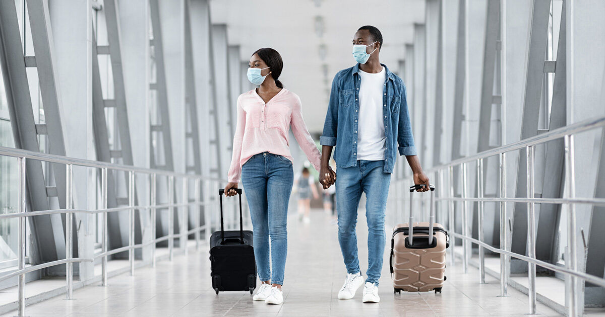 A young couple is seen walking side by side through a modern airport terminal, each pulling a rolling suitcase. Both individuals are casually dressed and wearing face masks, suggesting a travel scenario during a health-conscious period. The setting features bright natural light, glass walls, and a spacious corridor, emphasizing a contemporary travel environment.