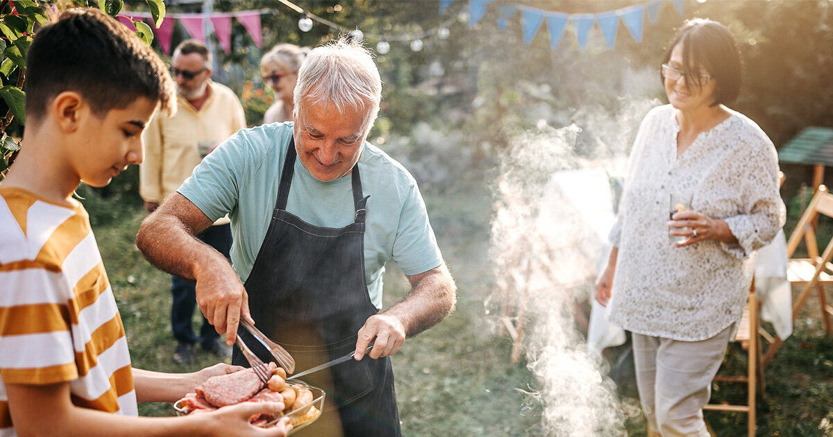 An older man in an apron slices grilled meat outdoors while a young person holds a plate. The scene takes place in a sunlit backyard with smoke rising from the grill and festive bunting in the background. Other adults are present, creating a relaxed and social atmosphere. The image captures a casual summer gathering with natural light and greenery.