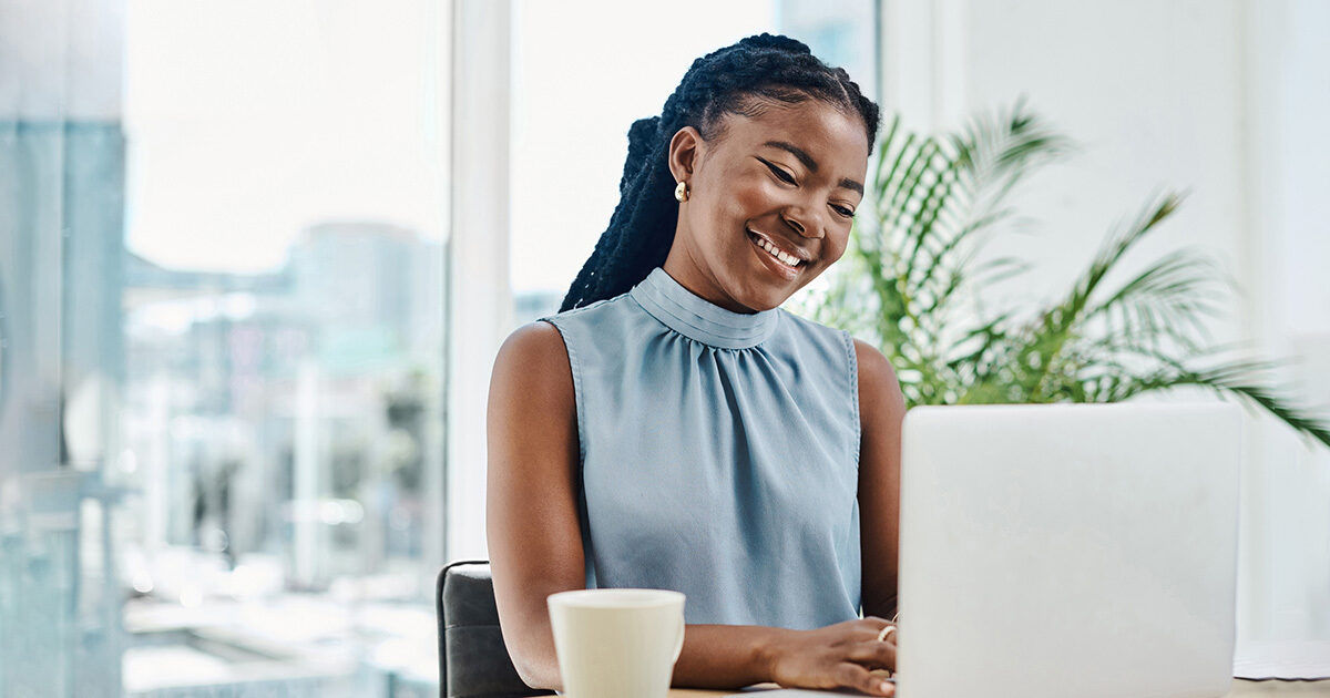 A woman sits at a desk with a white laptop and a coffee cup in a modern, sunlit office. Large windows and a green plant create a fresh, professional atmosphere. She wears a sleeveless light blue blouse and appears focused on her work. The setting suggests a contemporary workspace with natural light and minimalistic decor.