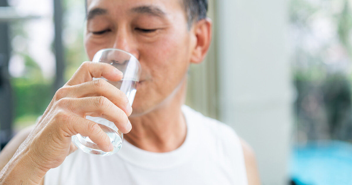 An older man is holding a clear glass of water close to his mouth. He is wearing a white sleeveless shirt and is seated in a bright indoor setting with natural light and greenery visible outside. The atmosphere is calm and healthy, emphasizing hydration and wellness. No visible text or numbers appear in the image.