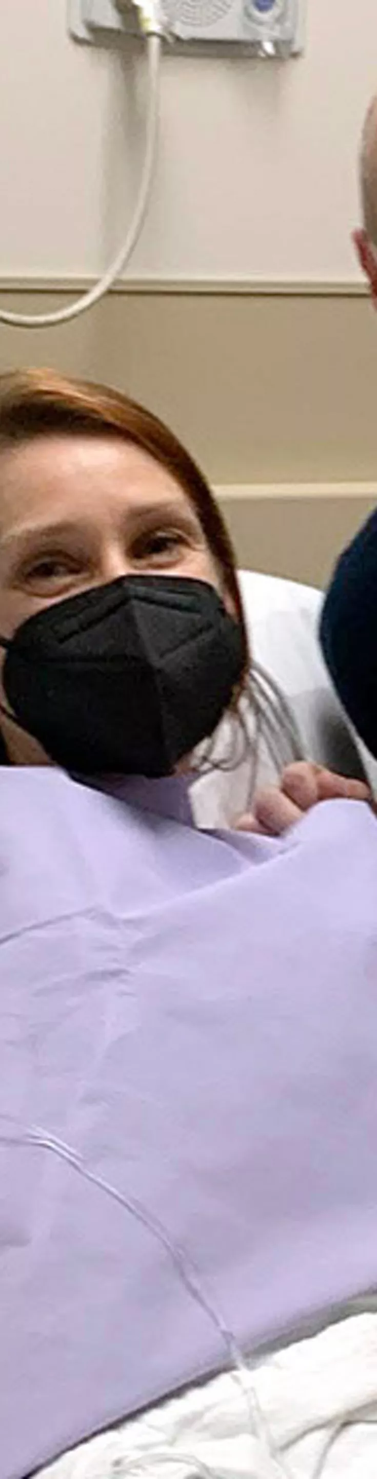 A patient is lying in a hospital bed, covered with a purple medical gown and blanket. Medical equipment and monitors are visible on the wall, indicating a clinical setting. An adult visitor sits beside the bed, showing support. The room contains standard hospital furnishings and medical devices. The atmosphere suggests care and recovery in a healthcare environment.