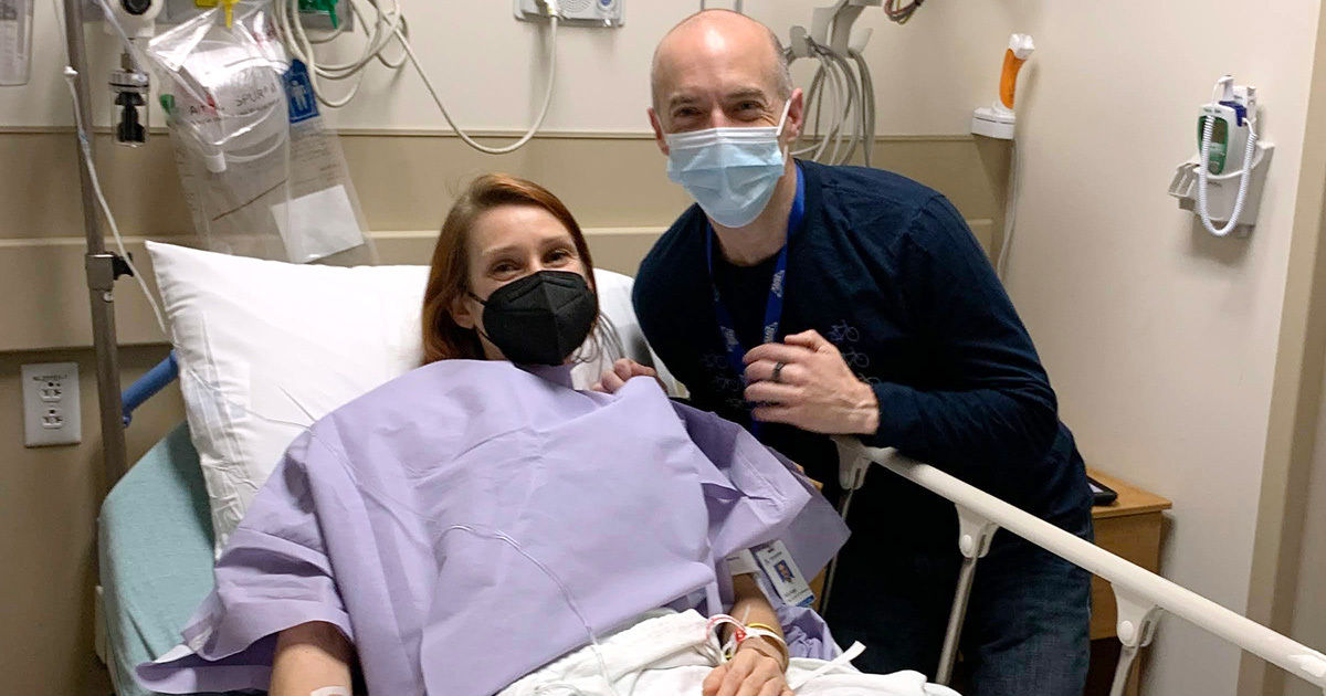 A patient is lying in a hospital bed, covered with a purple medical gown and blanket. Medical equipment and monitors are visible on the wall, indicating a clinical setting. An adult visitor sits beside the bed, showing support. The room contains standard hospital furnishings and medical devices. The atmosphere suggests care and recovery in a healthcare environment.