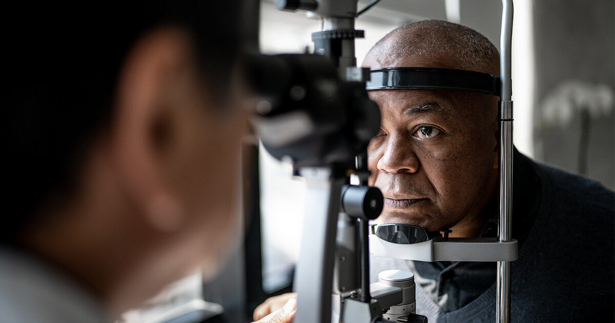 A person is seated at an ophthalmology clinic, having their eyes examined with a slit lamp device. The medical professional is positioned opposite the patient, focusing on the diagnostic procedure. The setting is clinical, with the slit lamp and examination equipment clearly visible. The lighting is neutral, emphasizing a professional healthcare environment.