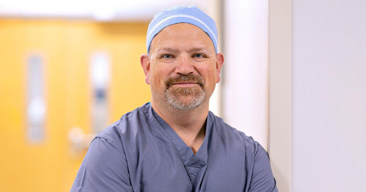 A healthcare worker is shown wearing blue scrubs and a surgical cap, standing with arms crossed in a brightly lit hospital corridor. The background features out-of-focus doors and neutral walls, suggesting a clinical environment. The overall mood is professional and calm, with a focus on the medical setting.