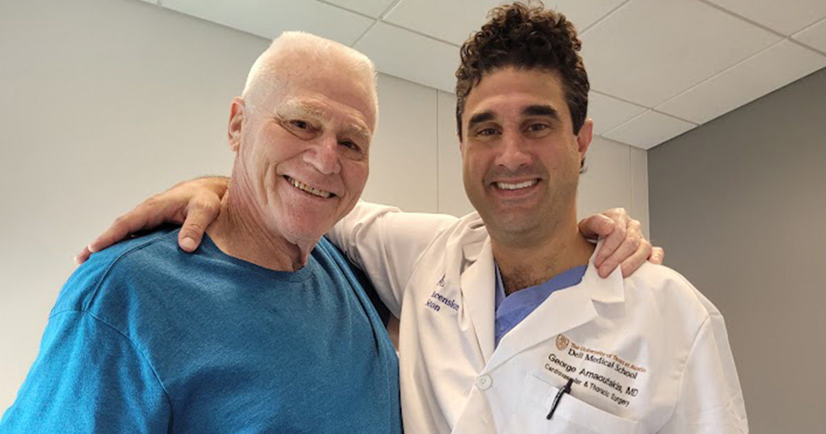 A medical professional in a white coat stands with an older adult male in a blue shirt. The setting appears to be a modern clinic or hospital room with neutral walls and a tiled ceiling. The doctor's coat displays visible text including 'MD' and 'Orthopaedic Surgery,' along with a name and institution. Both individuals are standing close, suggesting a supportive or friendly interaction.