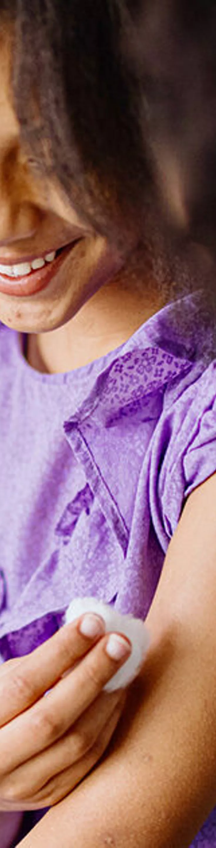 A young child wearing a purple shirt is shown after receiving a vaccination, with a bandage on their upper arm. A healthcare professional in blue gloves is gently cleaning the area. The background includes a blurred office or clinic environment with neutral tones. The scene conveys a sense of care and health awareness.