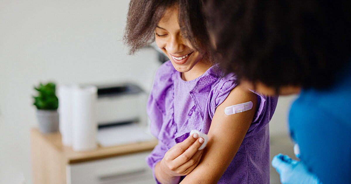A young child wearing a purple shirt is shown after receiving a vaccination, with a bandage on their upper arm. A healthcare professional in blue gloves is gently cleaning the area. The background includes a blurred office or clinic environment with neutral tones. The scene conveys a sense of care and health awareness.