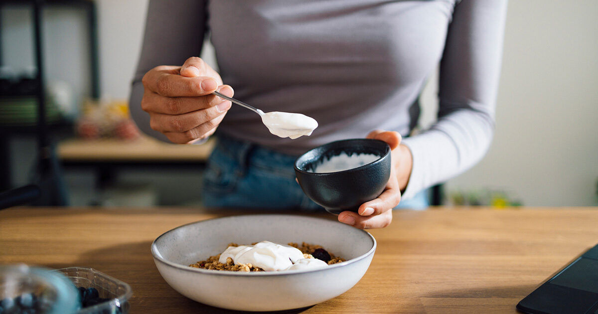 A woman is seen spooning yogurt from a small bowl onto a breakfast granola bowl. The setting appears to be a modern kitchen with a wooden countertop. The focus is on the hands and the food, with a casual and natural atmosphere. The color palette is neutral with soft lighting. No visible text or numbers are present in the image.