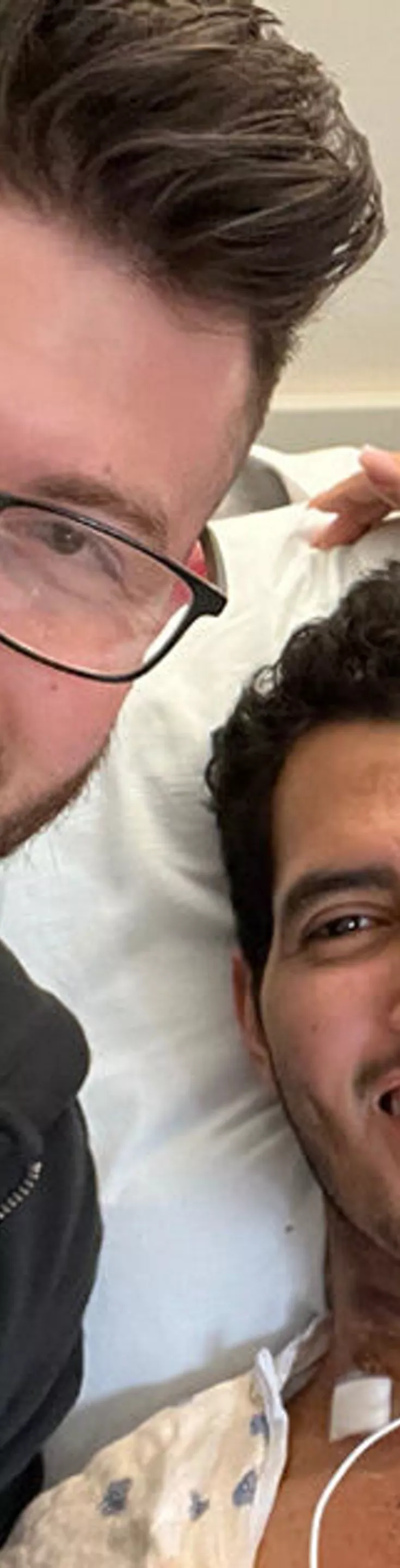 A patient lies in a hospital bed connected to medical monitoring wires, accompanied by two visitors. The setting is a clinical hospital room with visible medical equipment and emergency indicators on the wall. The visitors appear to be showing support and care, creating a compassionate and concerned atmosphere. The image conveys themes of recovery, support, and healthcare.