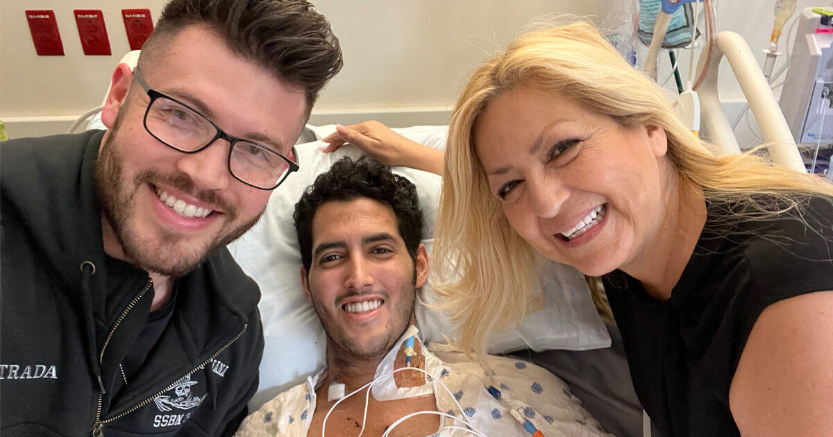 A patient lies in a hospital bed connected to medical monitoring wires, accompanied by two visitors. The setting is a clinical hospital room with visible medical equipment and emergency indicators on the wall. The visitors appear to be showing support and care, creating a compassionate and concerned atmosphere. The image conveys themes of recovery, support, and healthcare.