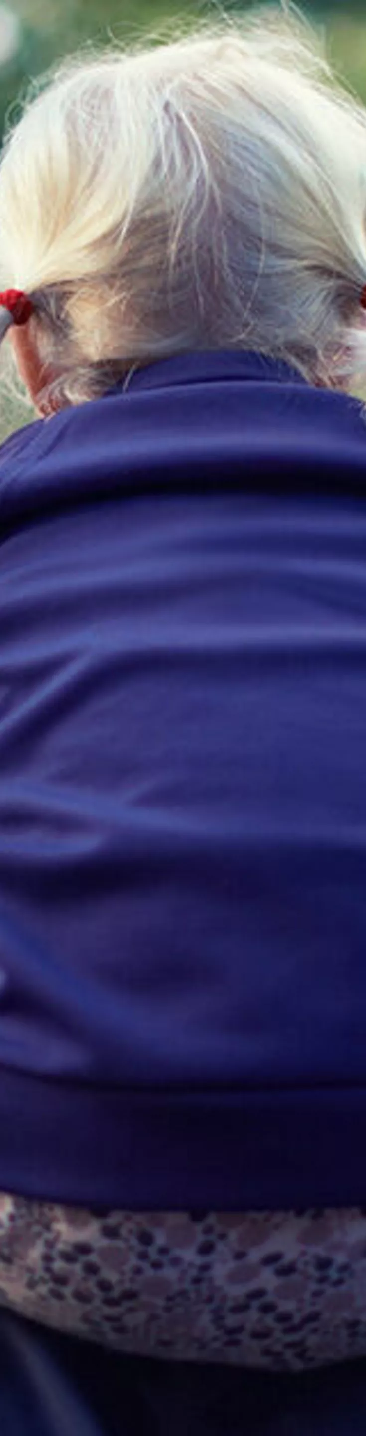 A young child with blonde hair in pigtails sits on an adult's shoulders, facing away from the camera. The setting appears to be an outdoor gathering or festival, with blurred lights and people in the background. The child is wearing a dark blue sweatshirt, and the overall mood is casual and joyful. The image is softly lit, with a cool color palette and a sense of lively atmosphere.