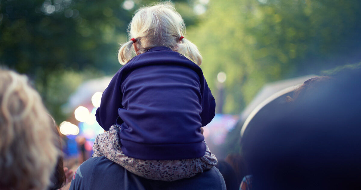 A young child with blonde hair in pigtails sits on an adult's shoulders, facing away from the camera. The setting appears to be an outdoor gathering or festival, with blurred lights and people in the background. The child is wearing a dark blue sweatshirt, and the overall mood is casual and joyful. The image is softly lit, with a cool color palette and a sense of lively atmosphere.