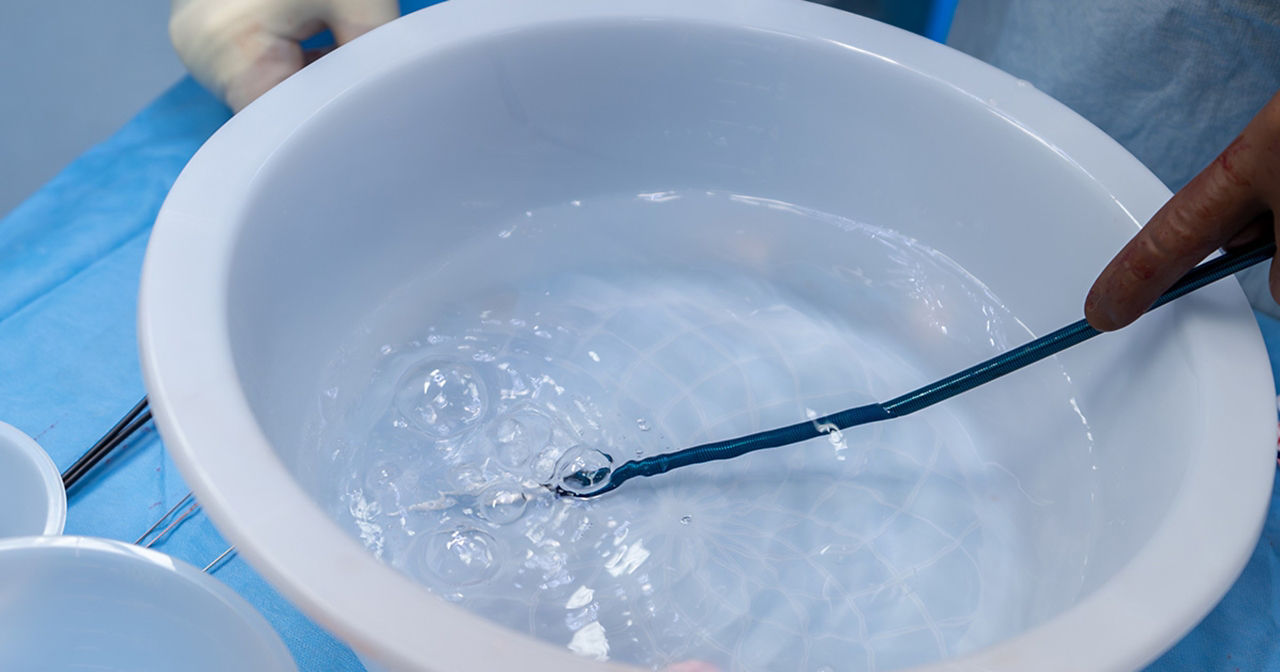 Surgical tool being rinsed in a sterile bowl of water during a medical procedure, with gloved hands and instruments visible nearby.