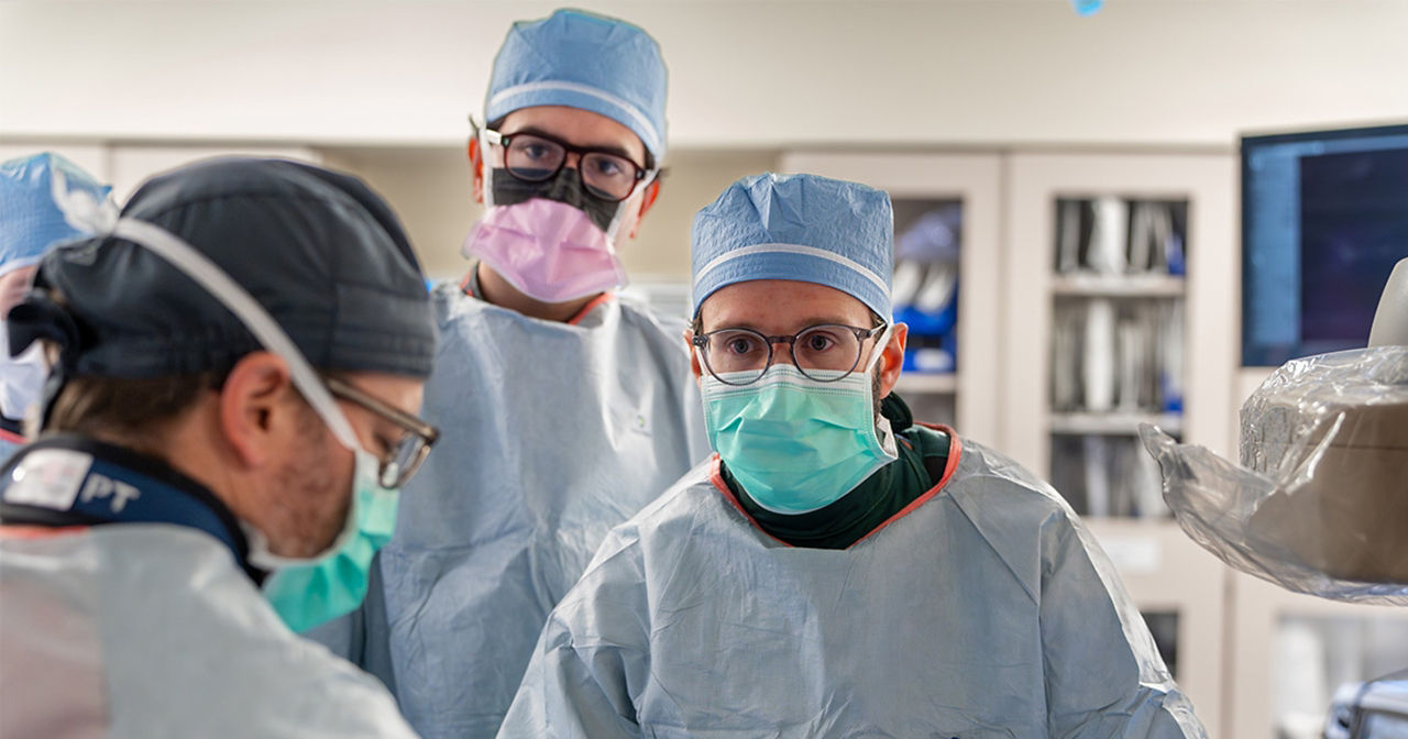 Three surgeons in scrubs, masks, and surgical caps performing a procedure in an operating room, with one surgeon focused on the camera and others concentrating on the patient.