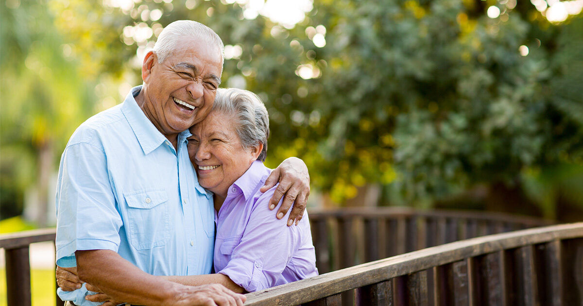 An elderly couple stands closely together, embracing on a wooden bridge in a lush, green park. The man wears a light blue shirt while the woman is dressed in lavender. Sunlight filters through the trees, creating a warm and peaceful atmosphere. The scene conveys affection and tranquility in an outdoor setting.