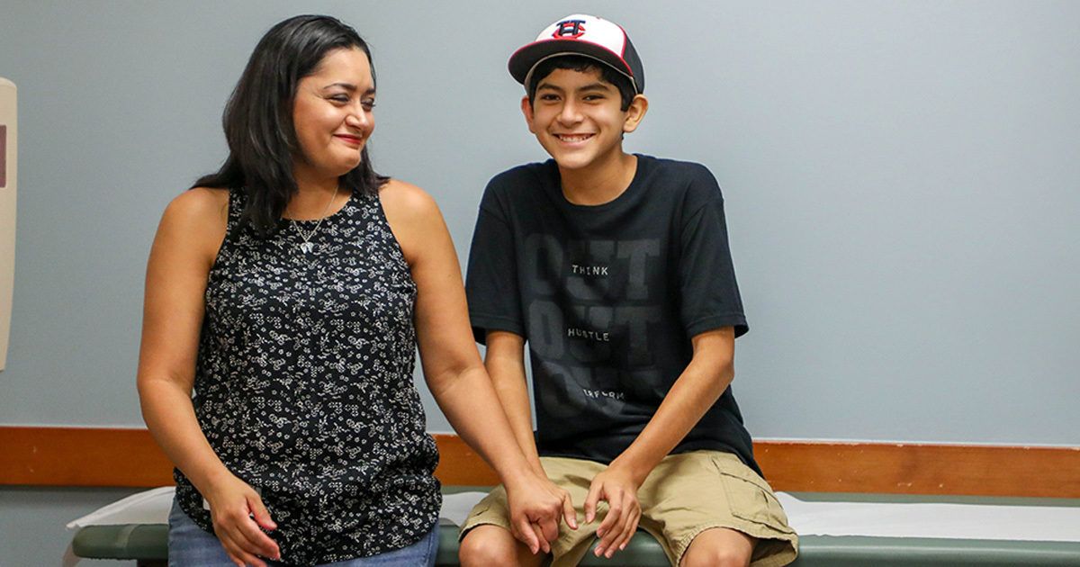 A woman and a teenage boy are seated side by side on an examination table in a clinical setting. The woman is wearing a sleeveless patterned top and the boy is dressed in a black t-shirt, khaki shorts, and a baseball cap. They are holding hands, suggesting support or comfort, with a neutral wall and medical equipment visible in the background. The overall mood is calm and supportive.