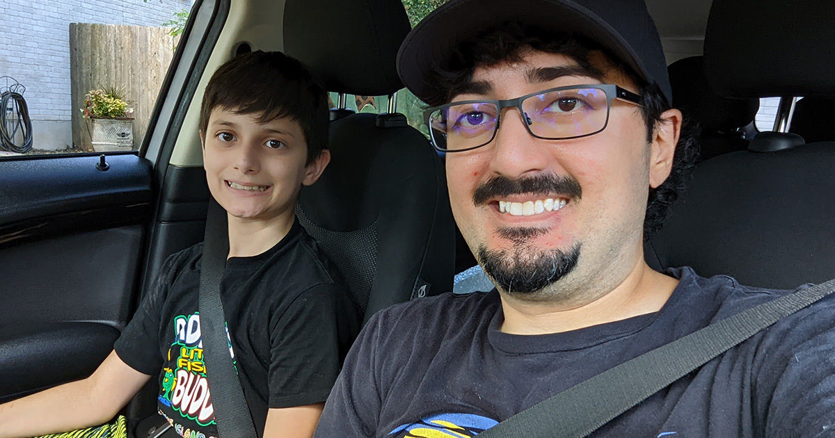 Two individuals are seated in the front seats of a car, both wearing seatbelts. The setting appears to be a parked vehicle in a residential area, with greenery and a brick wall visible outside the window. The interior is well-lit by natural daylight, and both people are dressed casually in t-shirts. The overall mood is relaxed and everyday.