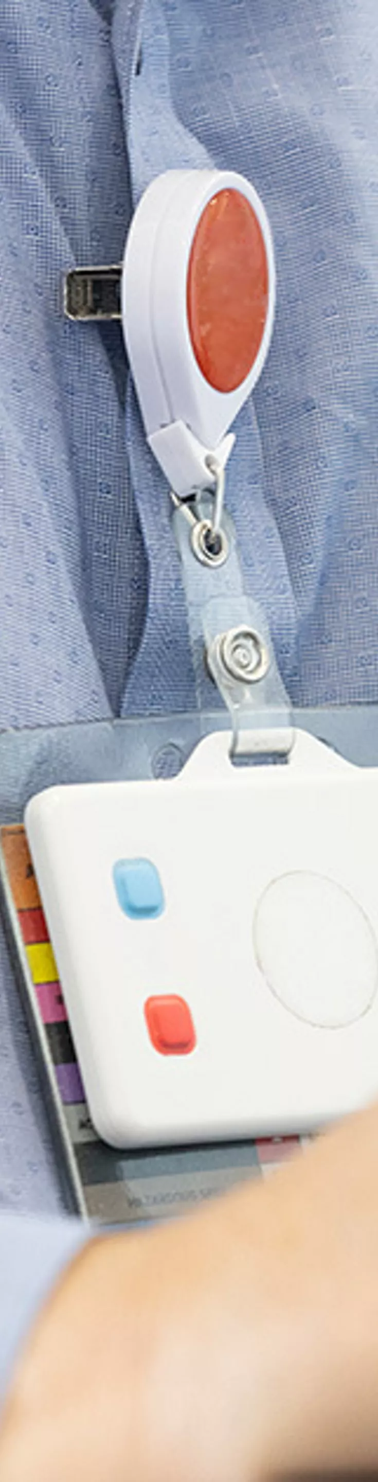 A person in a blue button-up shirt is working on a laptop, with focus on their hands and torso. An ID badge with a retractable clip and a pen in the shirt pocket are clearly visible. The setting appears to be an office environment, suggesting a professional or business context. 