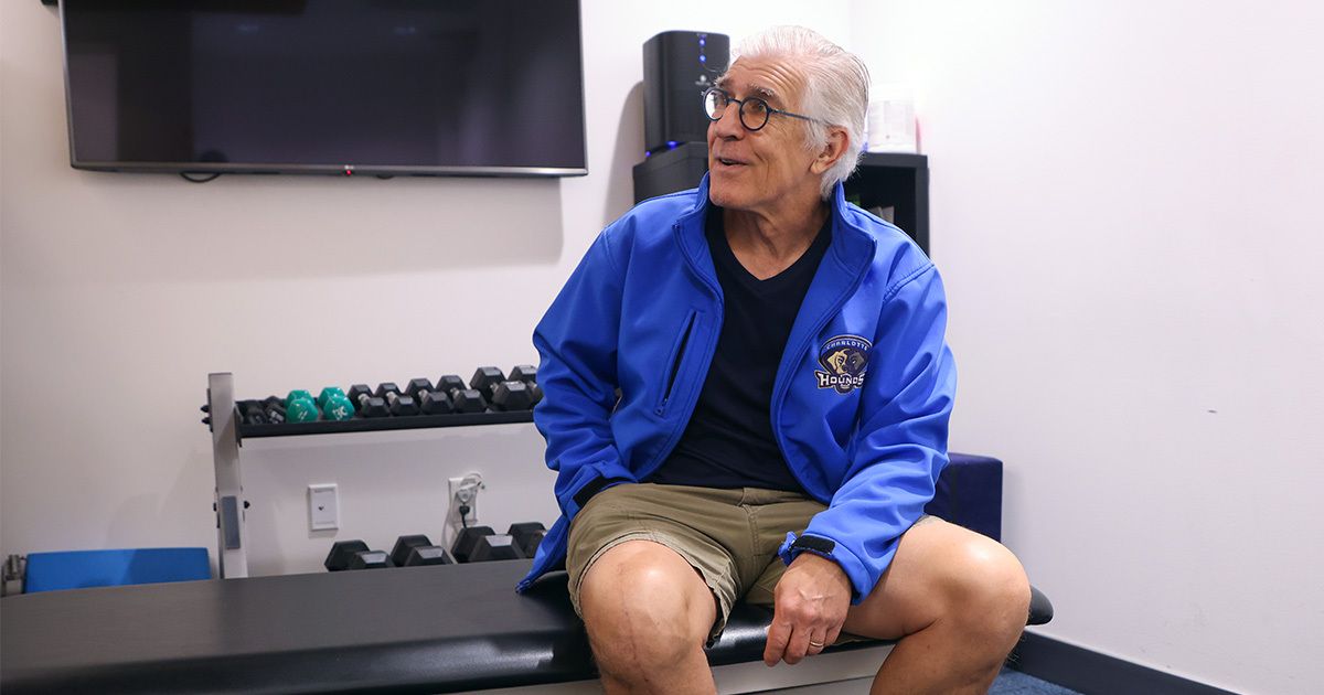 An older man with gray hair sits on an examination table in a clinical setting, wearing a blue jacket and khaki shorts. The room features a rack of dumbbells, a television mounted on the wall, and a clean, minimalist design. The man appears relaxed, possibly waiting for a physical therapy session or medical consultation. The environment is well-lit with neutral tones and functional decor.