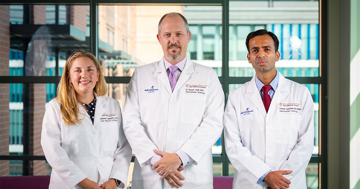 Three adults stand together indoors, each wearing a white lab coat and formal attire. The setting appears to be a modern medical or hospital environment, with large windows and urban architecture visible in the background. The group includes both men and women, suggesting a diverse medical team. Some embroidered text is visible on the lab coats, indicating professional titles or affiliations.