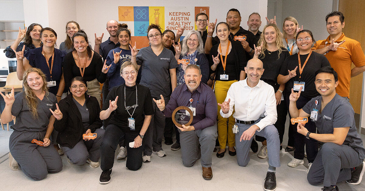 A large group of adults, many in medical scrubs and ID badges, pose together indoors. The setting appears to be a healthcare or office environment, with posters and signage visible in the background, including one that reads 'KEEPING AUSTIN HEALTHY SINCE 2017.' Several people hold small items and make hand gestures, suggesting a celebratory or team-building event. The mood is upbeat and collaborative.