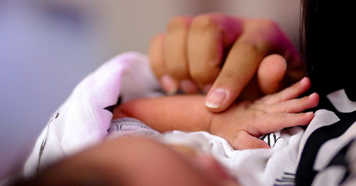 A gentle close-up captures an adult hand tenderly holding a newborn baby's tiny hand. The scene is intimate, with soft focus and warm lighting emphasizing the emotional bond. The baby is wrapped in a light-colored blanket, and only part of the adult's face and hand are visible. The overall mood is nurturing and serene.