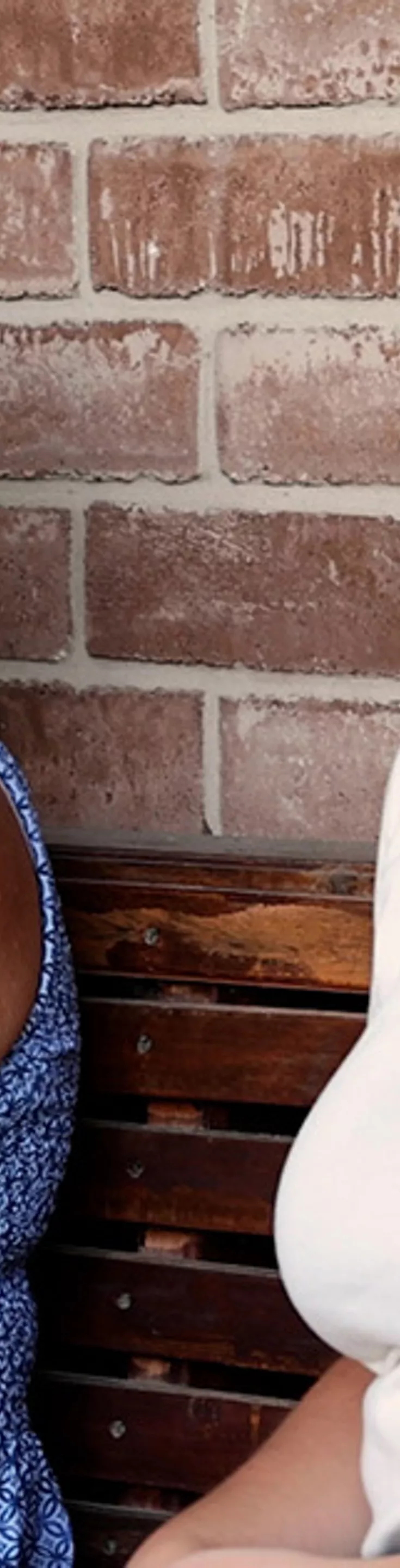 An older woman in a blue patterned dress and a younger woman in a white t-shirt are seated side by side on a wooden bench. The setting is an outdoor porch with a brick wall and a potted plant visible in the background. The mood is relaxed and casual, with natural daylight illuminating the scene.