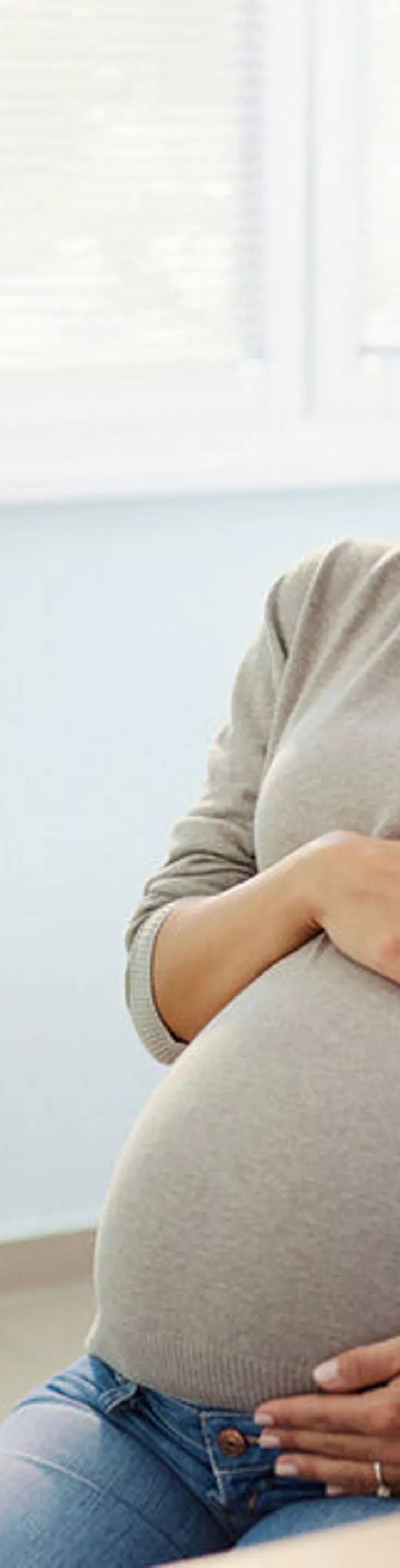 A pregnant woman sits in a medical office, holding her belly while speaking with a healthcare professional. The setting is a bright, modern clinic with medical equipment and documents visible. The healthcare provider appears to be explaining or discussing something important. The overall mood is calm and supportive, with neutral and soft color tones. No visible text or numbers are present in the image.
