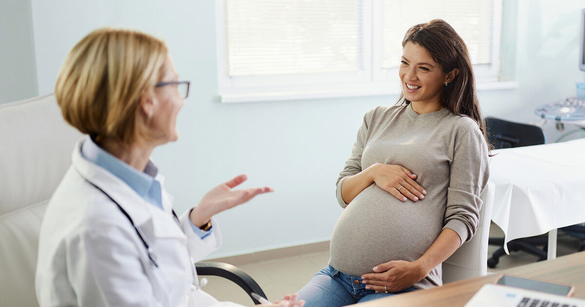 A pregnant woman sits in a medical office, holding her belly while speaking with a healthcare professional. The setting is a bright, modern clinic with medical equipment and documents visible. The healthcare provider appears to be explaining or discussing something important. The overall mood is calm and supportive, with neutral and soft color tones. No visible text or numbers are present in the image.