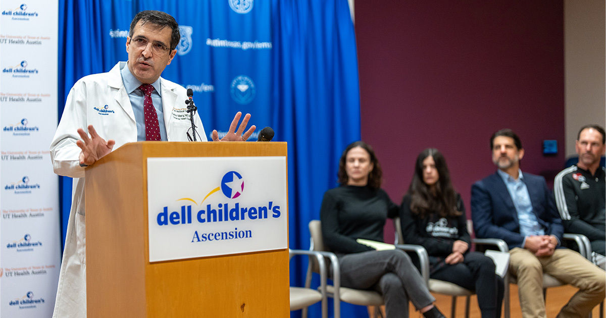 A medical professional in a white coat addresses an audience from a podium at a Dell Children's Ascension event. The setting is a formal indoor press conference or panel, with several seated adults in business and casual attire. The background features a blue curtain with visible Dell Children's Ascension branding and a maroon wall. The atmosphere is professional and informative.