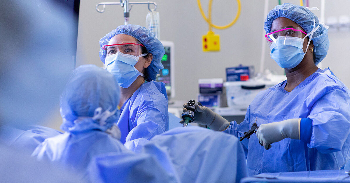 Medical professionals in blue surgical gowns and caps are conducting a laparoscopic surgery in a modern operating room. The scene includes advanced surgical instruments and visible IV equipment. The background shows medical supplies and a yellow wall-mounted device. The environment is brightly lit and sterile, emphasizing a clinical and professional atmosphere.