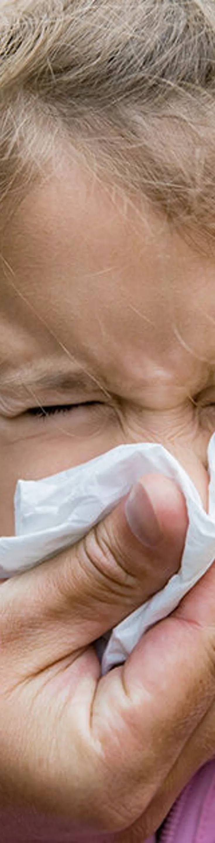 An adult assists a young child by holding a tissue to the child's nose. The scene takes place outdoors, with autumn foliage visible in the background. The child is wearing a light pink jacket, and the adult's hand is visible, suggesting care and attention. The overall mood is nurturing and candid, with natural lighting and earthy tones.