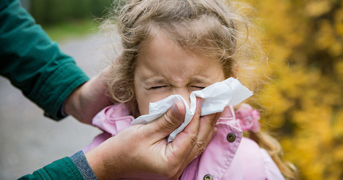 An adult assists a young child by holding a tissue to the child's nose. The scene takes place outdoors, with autumn foliage visible in the background. The child is wearing a light pink jacket, and the adult's hand is visible, suggesting care and attention. The overall mood is nurturing and candid, with natural lighting and earthy tones.